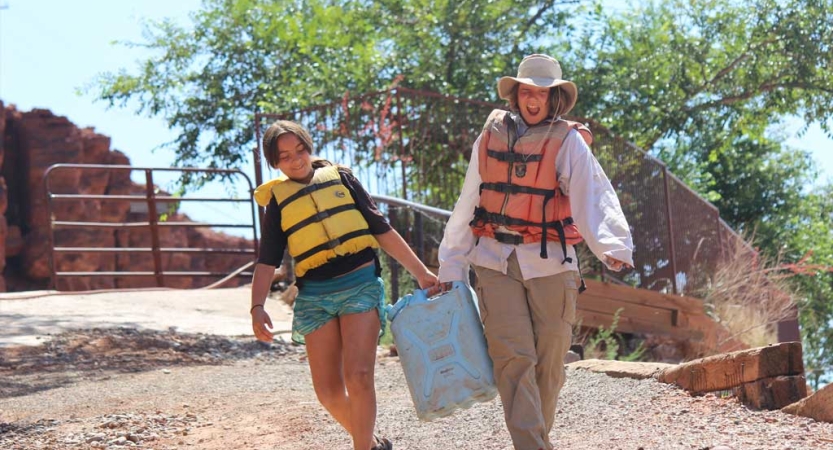 Two people wearing life jackets carry a large jug together